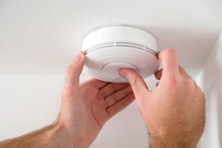 A smoke alarm fitted to a ceiling and a mans hands pushing the smoke alarm test button.