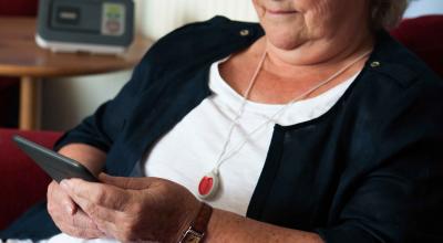 An elderly woman sitting in an armchair using her tablet wearing a community alarm pendant. The pendent is on a small chain around her neck. The pendant as a large red button.