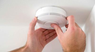 A smoke alarm fitted to a ceiling and a mans hands pushing the smoke alarm test button.